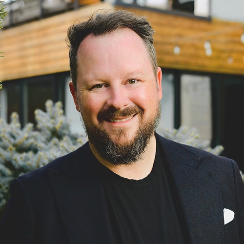 A man with a beard smiling in front of a house.